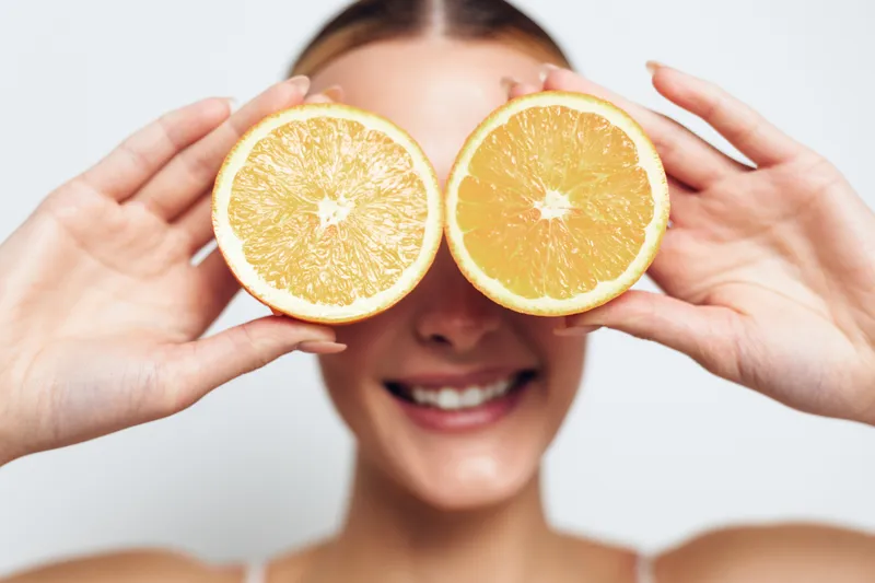 Smiling female with brunette hair holds two orange halves in front of her eyes, bright clean background.