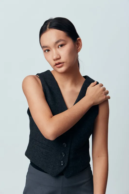 Young woman with dark hair and natural makeup wearing a black vest and gray pants posing with hand on shoulder against a light gray background in studio portrait.