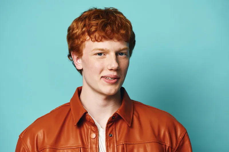 Portrait of young man with red hair wearing brown leather jacket looking at camera with slight smile against blue background in studio lighting. Casual confident expression with natural skin tone.
