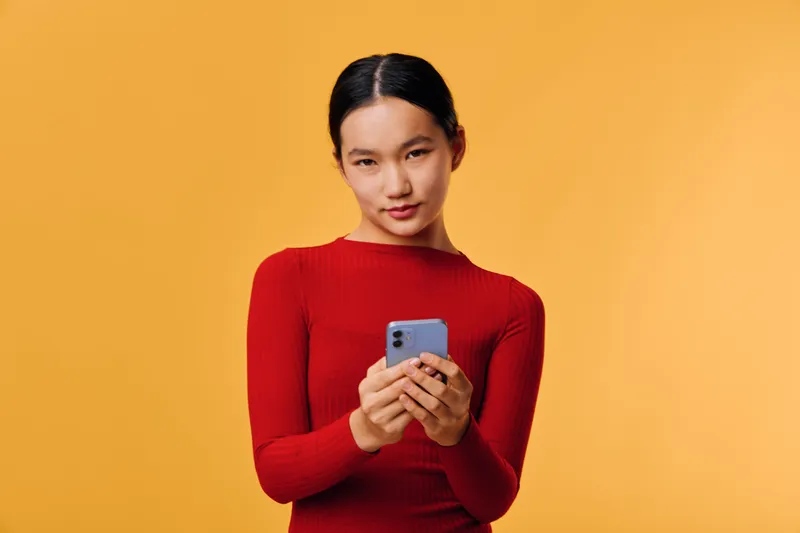 Young woman in red sweater holding smartphone and texting message with focus on screen against yellow background for communication and technology.