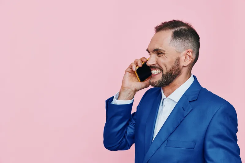 Professional man on a colored pink background smiles while talking on a phone, projecting confidence, approachability, and positive business energy for clients and colleagues.