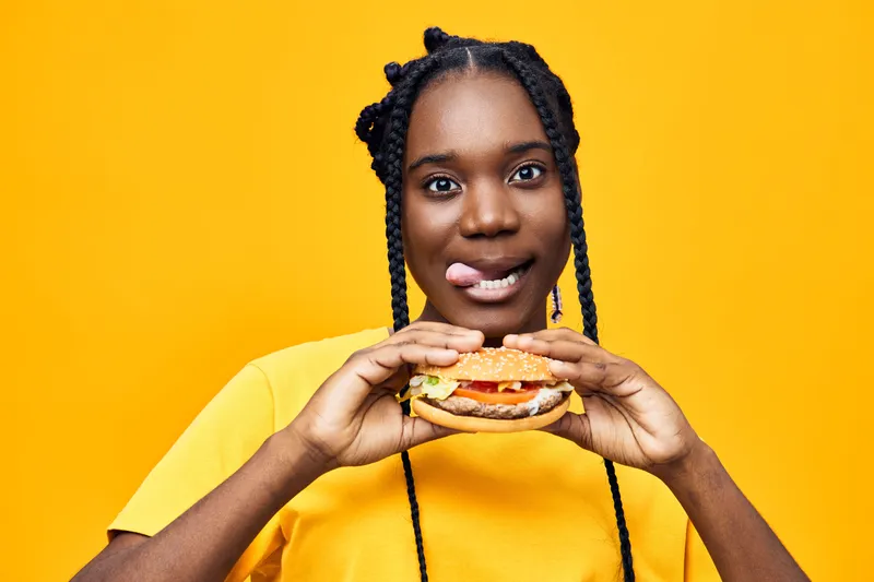 Young Black woman smiling and holding a delicious burger against a vibrant yellow background, showcasing joy and a love for fast food