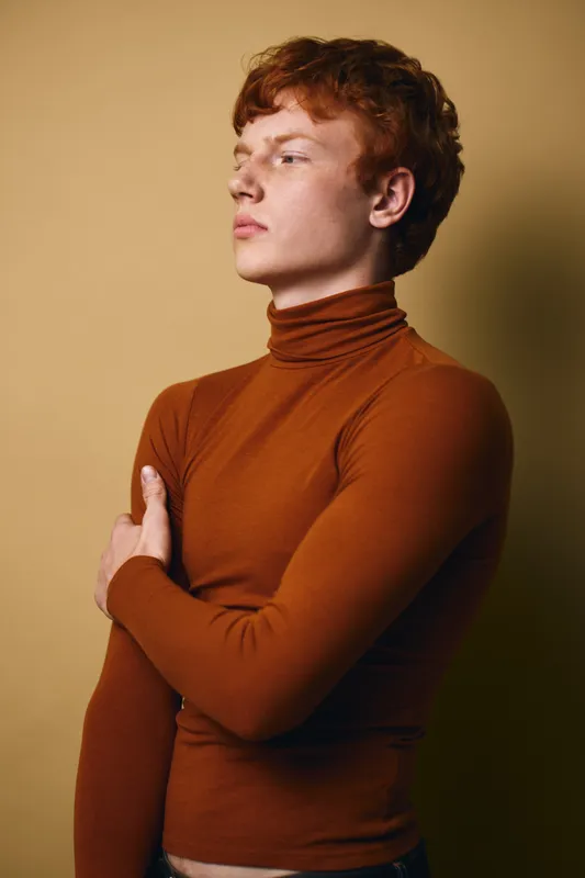 Redhead man with curly hair wearing brown turtleneck standing sideways against beige background looking away with thoughtful serious expression and folded arms.
