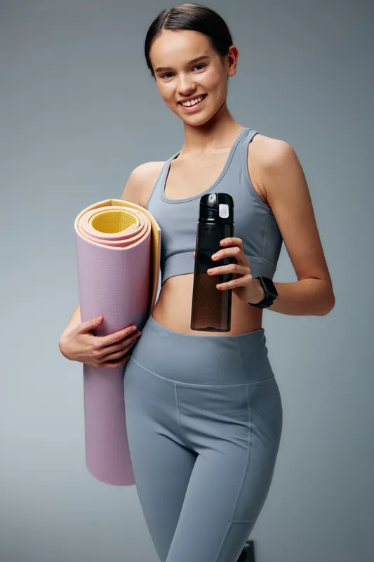Young woman in grey athletic wear holding a yoga mat and a water bottle, standing in a neutral studio. Fitness lifestyle, workout routine and yoga practice ready for training.