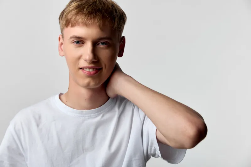 Young man in a white tee adjusts his neck in a casual studio pose, relaxed and confident, clean background, natural expression, minimal fashion vibe and mood