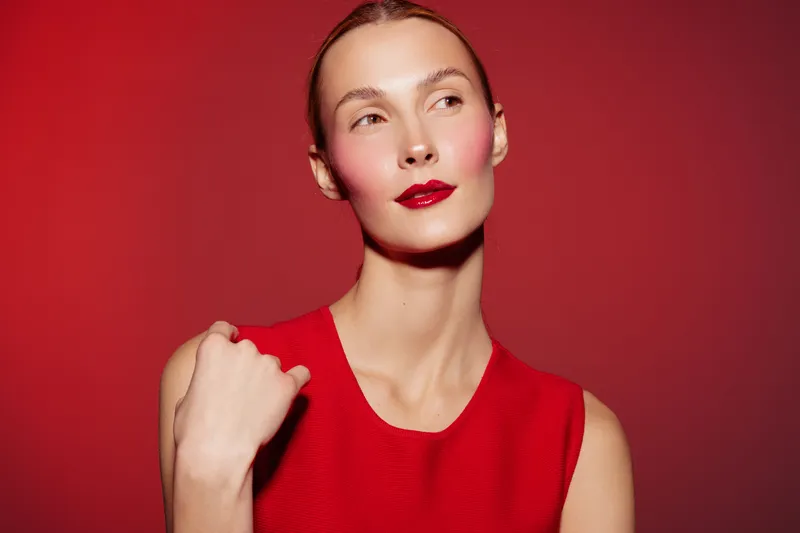 Elegant woman with red lipstick and red dress poses against red background. Studio portrait with smooth skin and confident expression. Concept of fashion, beauty, and style.
