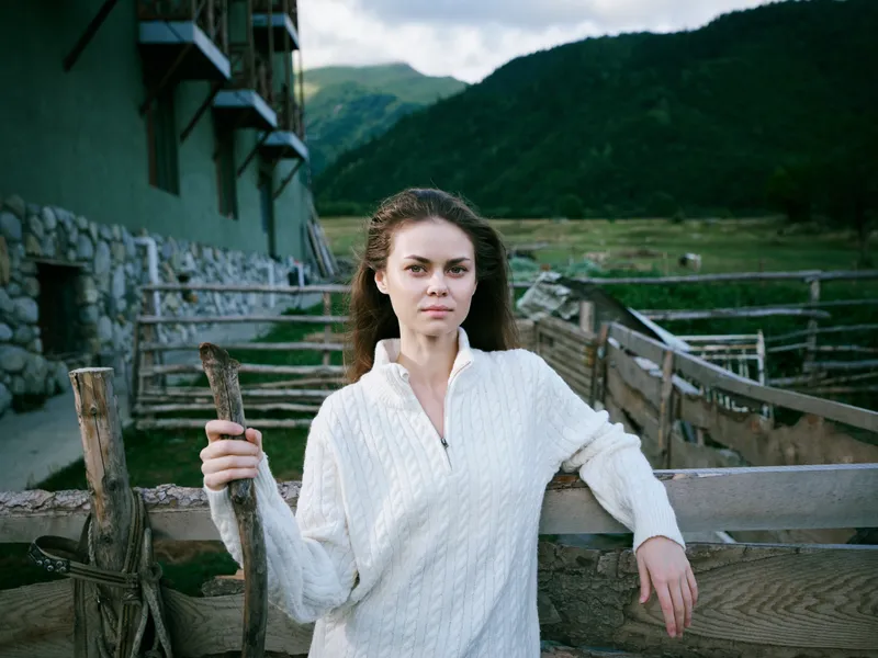 Woman in white knit sweater leans on a wooden fence in a farm setting, with stone buildings and distant mountains, capturing calm rural life and outdoor country style