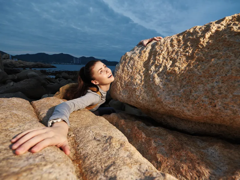 A young woman embracing a large rock formation on a coastal landscape, showcasing determination and adventure in a serene setting with soft lighting