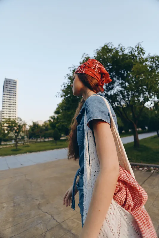 lifestyle woman in modern boho-western denim street style walks outdoors with red bandana and crochet bag, captured in warm film color tones under clear sky