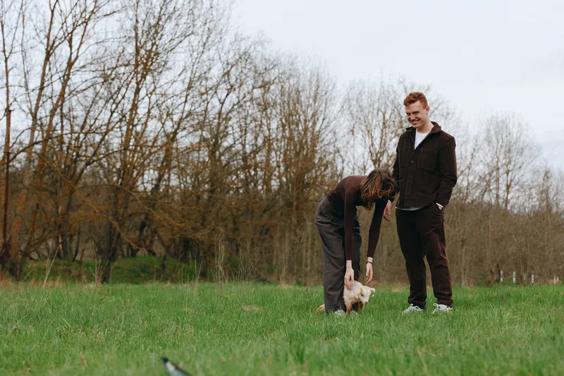 A pair of men outdoors in a field of grass interact with a goat kid, capturing a warm, candid moment of animal companionship and rural life in daylight.