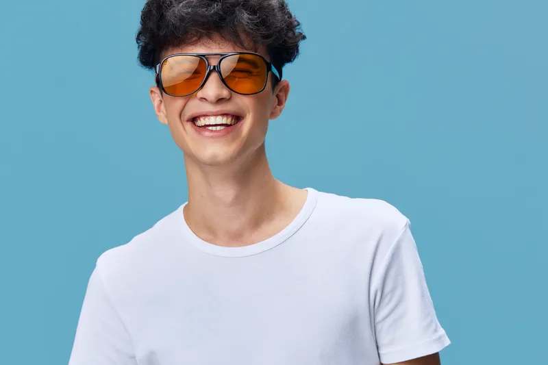 Smiling man wearing yellow sunglasses and white t-shirt, showing happiness and youthfulness against a bright blue background with casual summer style.