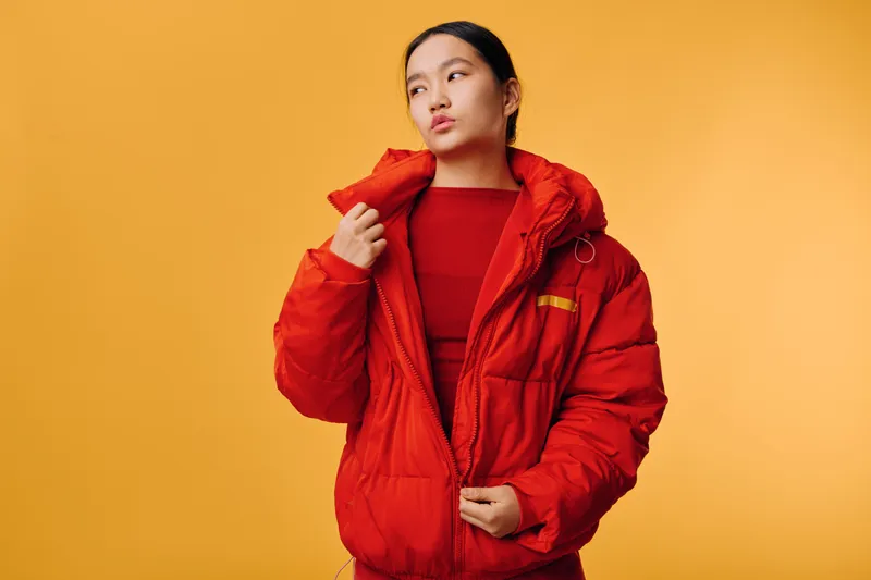 Woman wearing red puffer jacket looking to side on yellow background. Casual clothing style with warm outerwear and confident expression in studio with solid color wall.