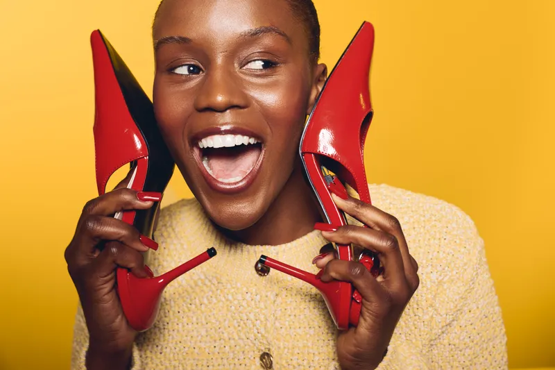 Happy African woman with dark skin holds red high heels near face, smiling widely. Yellow background and cozy beige sweater create warm, joyful fashion concept with vibrant color contrast.