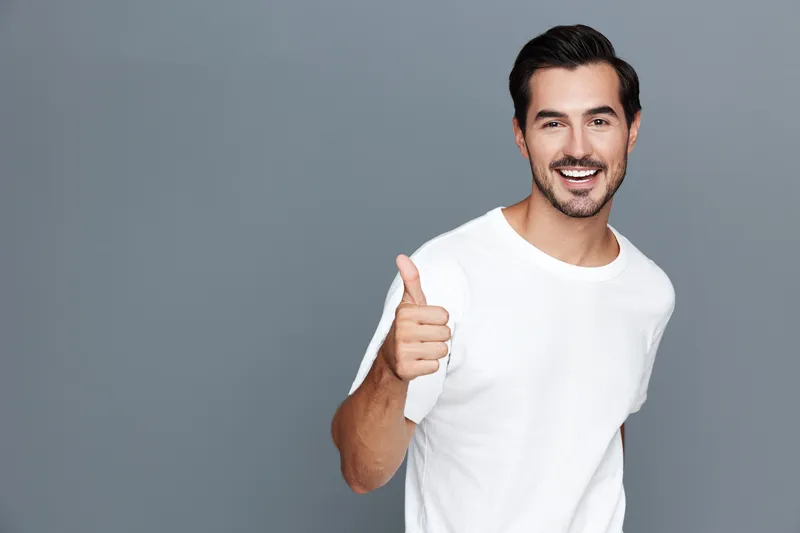Smiling man in a casual white t-shirt giving a thumbs up gesture against a grey background, exuding happiness and positivity in a portrait style.