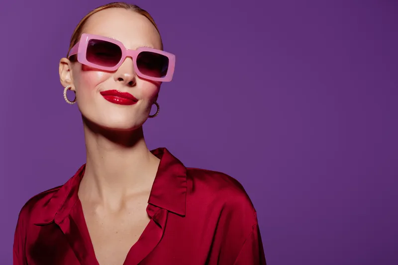 Stylish woman with pink sunglasses and red lipstick poses confidently against purple background. Fashion concept with bold colors and modern accessories for trendy look.