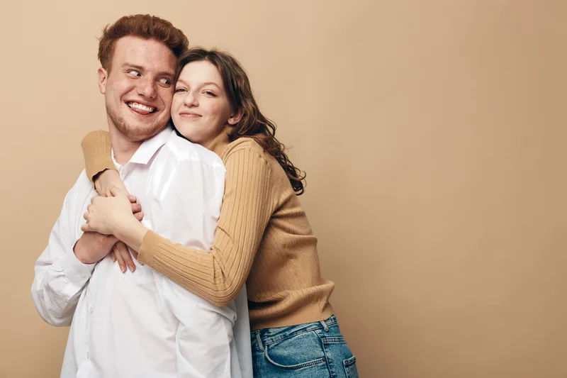 Happy young couple embraces and smiles together against beige background. Casual clothing and warm mood create concept of love, friendship, and relationship between man and woman.