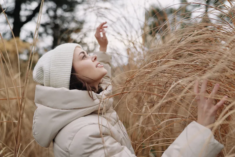 Woman in a winter coat and beanie among tall dried grass outdoors, enjoying the chilly air and nature in a peaceful winter landscape.