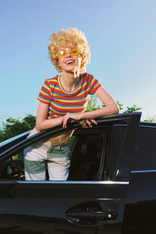 Playful woman outdoors, leaning from a sleek car window, striped top, retro style glow, sunlit colors, candid lifestyle moment with warm breeze and joyful smile