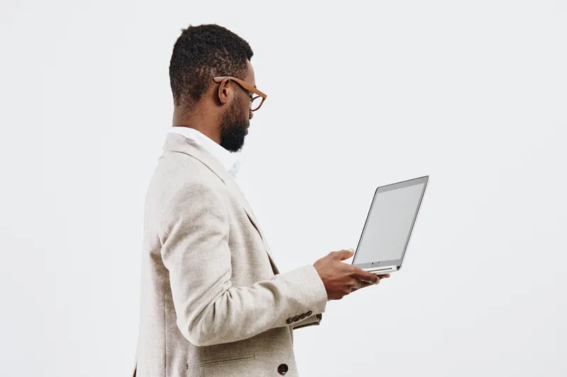 Stylish professional man with beard and glasses holding open laptop in hand, wearing light beige blazer on plain white background studio portrait. Business technology concept