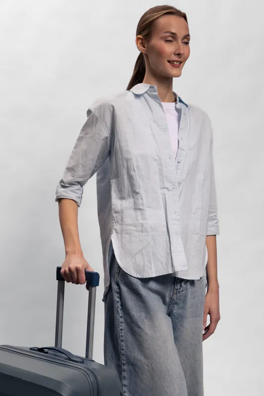 Young woman with suitcase stands against white background smiling slightly. Casual clothes and relaxed posture create concept of travel, vacation, and light packing.