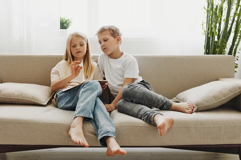 Two children sitting barefoot on beige couch using digital tablet together in bright living room with plants and natural light. Concept for technology, childhood, and family bonding.