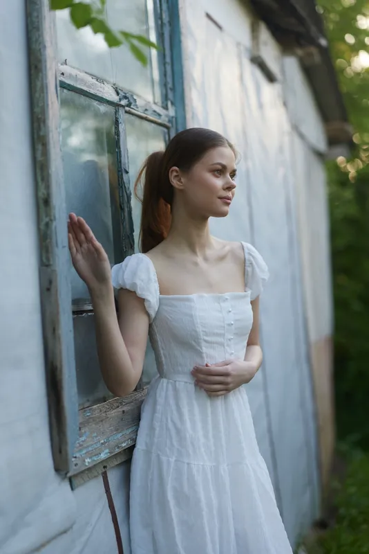 Young woman in a white summer dress standing by an old wooden window, expressing calm emotions and embracing the serenity of nature