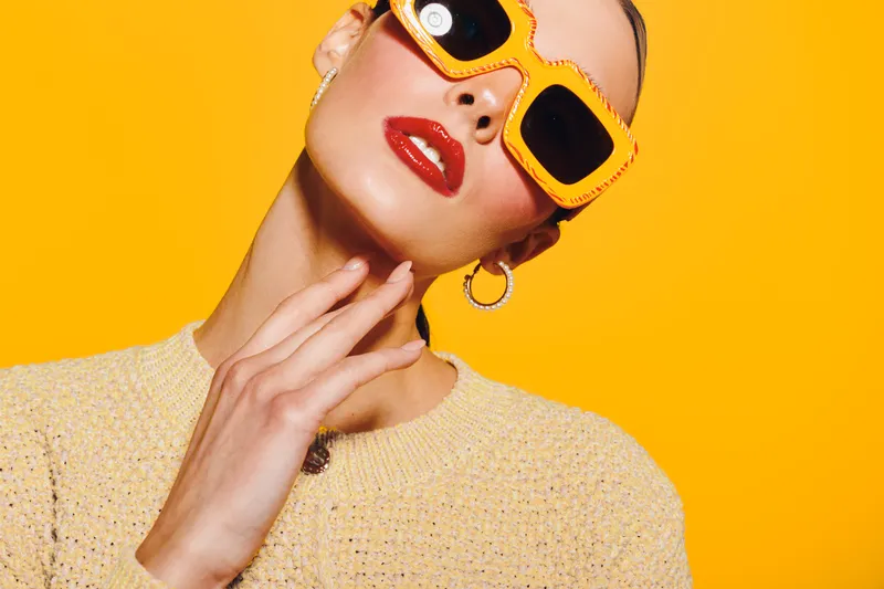 Stylish woman with bright orange sunglasses and red lipstick poses against yellow background. Fashion concept with trendy accessories and confident attitude in vibrant studio lighting.