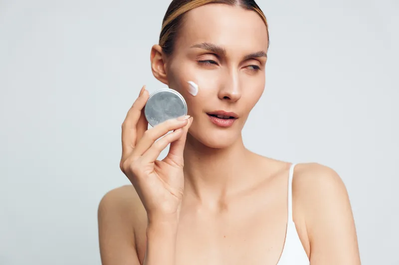 Young woman with bare shoulders applies moisturizing cream on her face holding cosmetic jar isolated on white background with natural skin care routine concept.