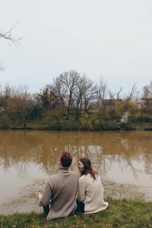 Young couple in cozy sweaters sits by the calm river enjoying a quiet moment together in early spring with bare trees and soft natural light around them