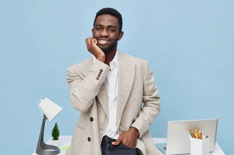 Confident smiling man with dark skin, casual beige blazer, white shirt, seated at modern workspace with laptop and desk lamp, on blue background. Creative, professional, positive, business, office, te