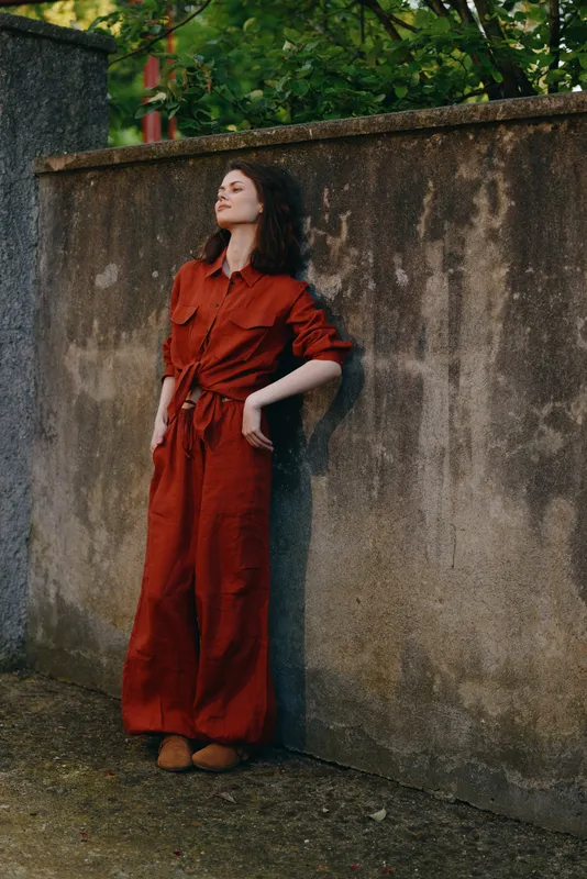 A stylish woman in a red jumpsuit stands against a weathered wall outdoors, posing confidently. Casual, urban lifestyle image exploring personal style and self-expression
