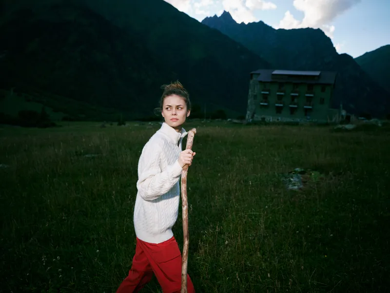 A woman wearing a white sweater walks with a wooden staff across a green field, with distant mountains and a hillside lodge, creating a rustic landscape of outdoor exploration and travel