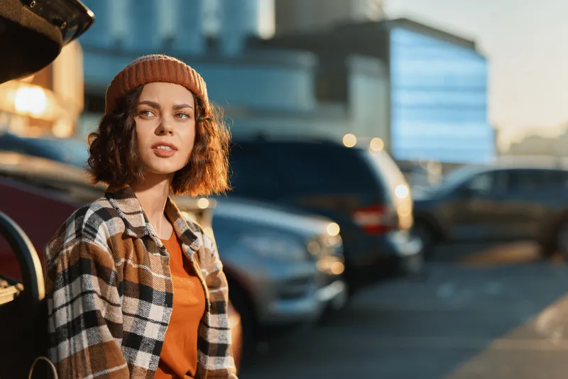 Woman portrait in urban parking wearing beanie and plaid jacket at golden hour, candid lifestyle shot with authenticity, golden hour glow, mindful living and emotional storytelling mood.