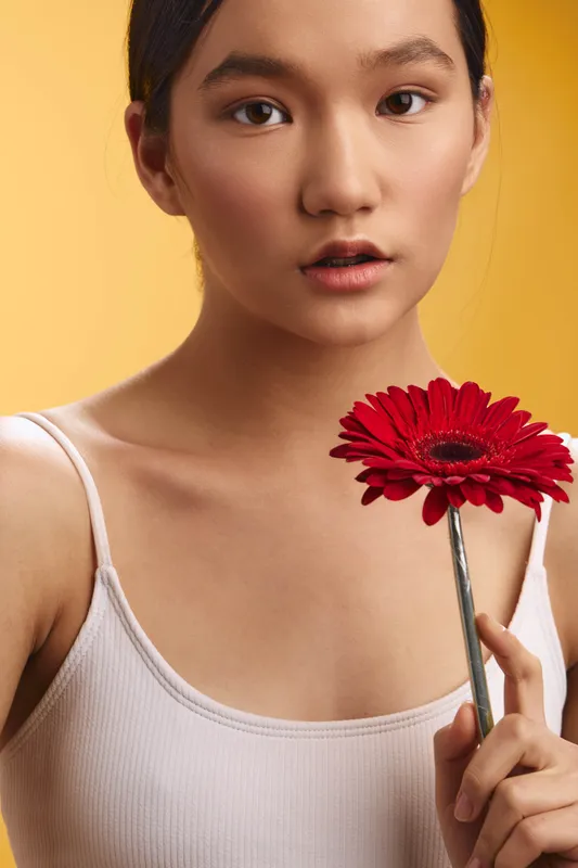 asian woman holds a red flower close to her face, soft expression, warm lighting, minimal setting, graceful pose that conveys calm beauty and thoughtful mood.