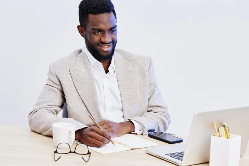 Professional smiling young African American man in beige blazer with white shirt working at desk with laptop, notebook, and stationery, studio indoor shot, modern workspace environment, positive mood,