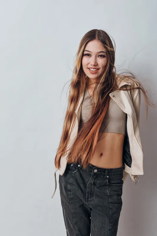 Young woman with long hair posing in casual fashion outfit standing against light background. Female model smiling with hands in pockets wearing crop top and jacket.