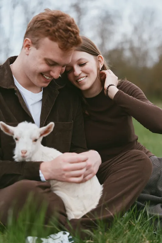 Young couple sitting on grass in casual clothes gently holding a baby goat while sharing a tender and joyful moment outdoors in a natural setting.