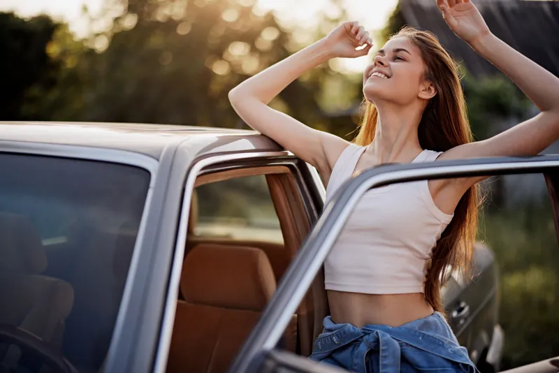 A young woman enjoying the sunlight outside a car, dressed in a casual summer outfit, representing freedom and joy in a natural setting.