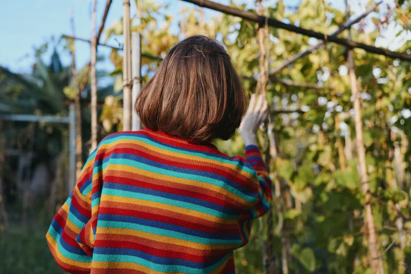 Woman in a striped sweater back view in a tropical outdoor garden, brown bob haircut reaching vines and touching leaves, casual outfit, relaxed mood and leisure lifestyle scene.