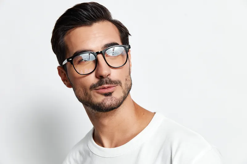 Stylish handsome young man in glasses poses confidently while looking away from the camera, showcasing a modern haircut and casual white sweater against a light background.