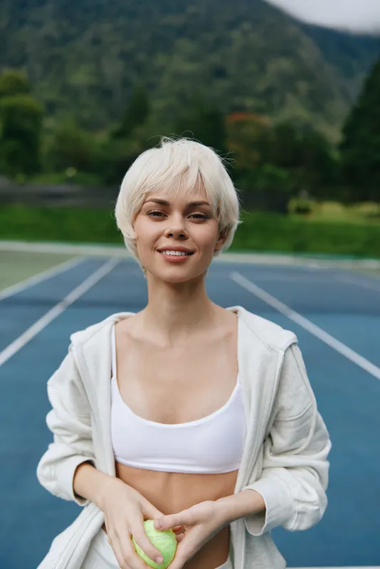 Young woman with short blonde hair in a sports bra and jacket, smiling confidently on a tennis court, exuding a vibrant and energetic atmosphere
