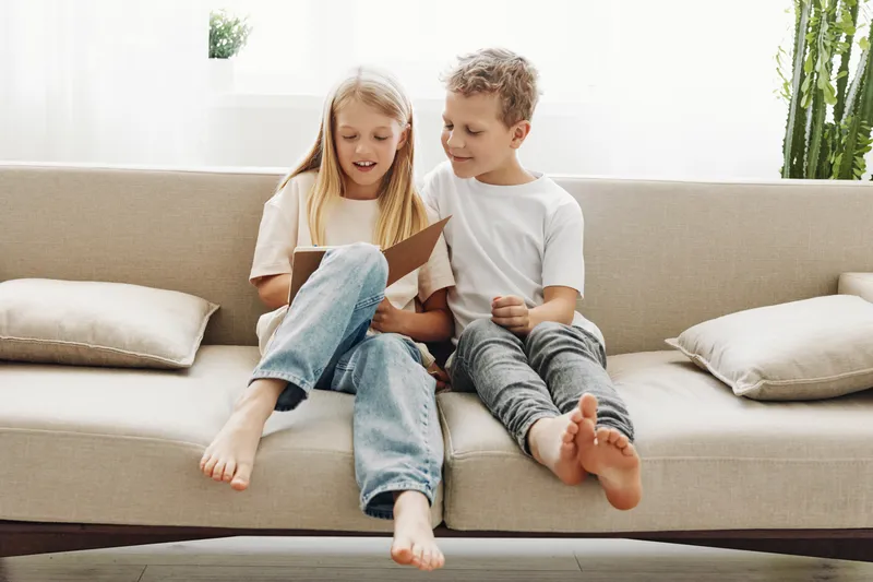 Two barefoot children sitting on a beige sofa reading a book together in a bright modern living room. Concept of childhood learning, friendship, and family bonding moments.