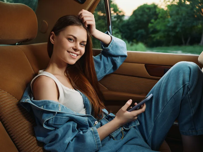 Smiling young woman in denim outfit enjoying a moment in a car, surrounded by greenery, with a casual and relaxed vibe, representing joy and comfort.
