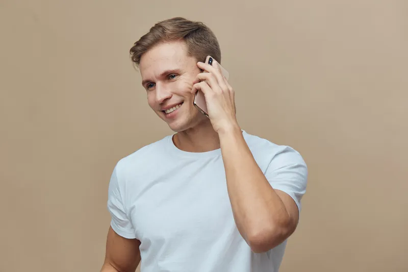 smiling young caucasian man talking on phone wearing white t-shirt isolated on plain beige background digital nomad remote work concept