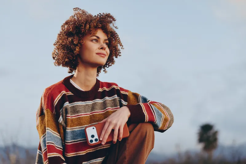 Woman lifestyle outdoors, curly hair, sweater, holding phone, soft clouds sky, relaxed smile, warm autumn colors, standing in open landscape, gentle breeze, casual street style