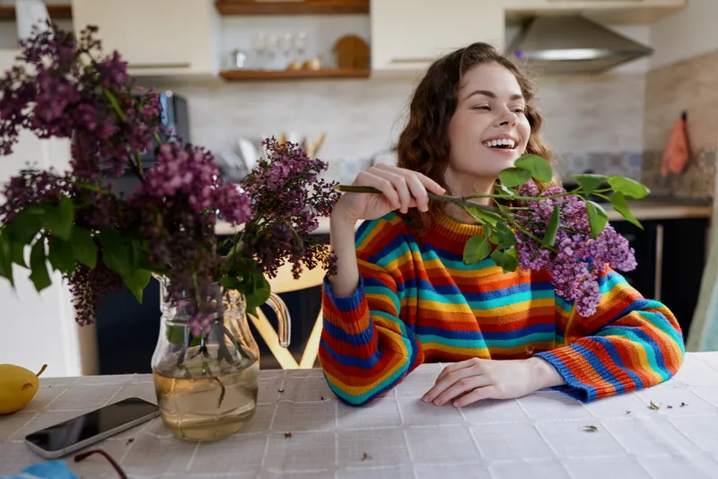 Joyful young woman indoors at a kitchen table smelling a lilac bouquet and smiling as natural light fills the cozy home