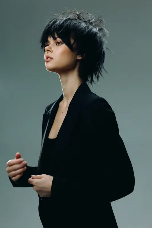 Stylish woman with short black hair wearing a black blazer, posing thoughtfully against a dark background, elegant fashion portrait.