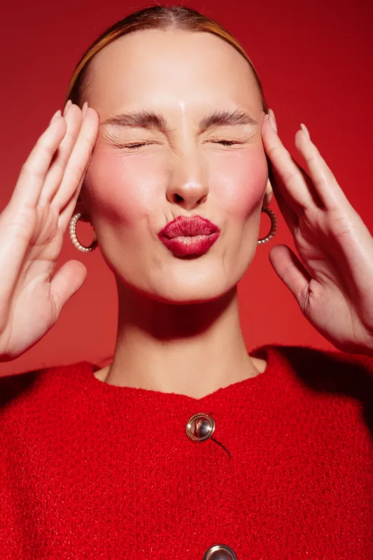 Close-up of a young woman with red lipstick and red sweater making a playful kiss face with closed eyes and hands near her temples. Bright red background creates a bold, vibrant mood.