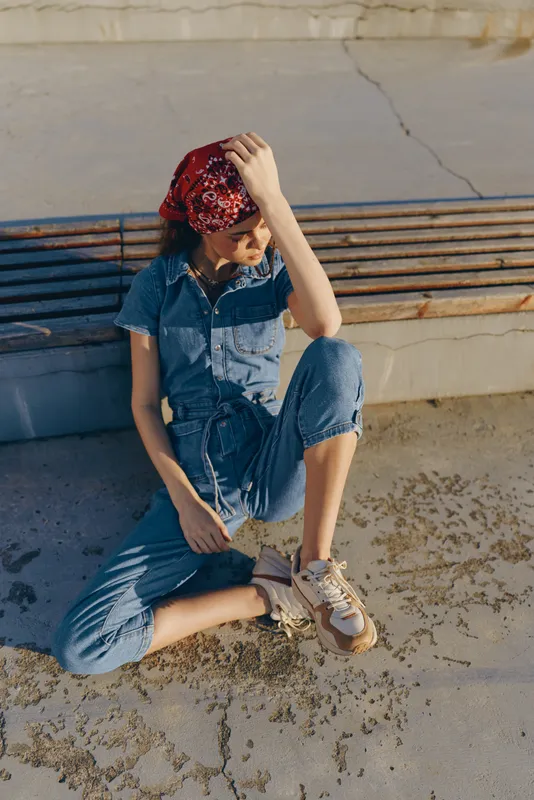 Lifestyle woman in modern boho-western denim street style wearing a red bandana and sneakers, sitting casually outdoors with warm film color tones.
