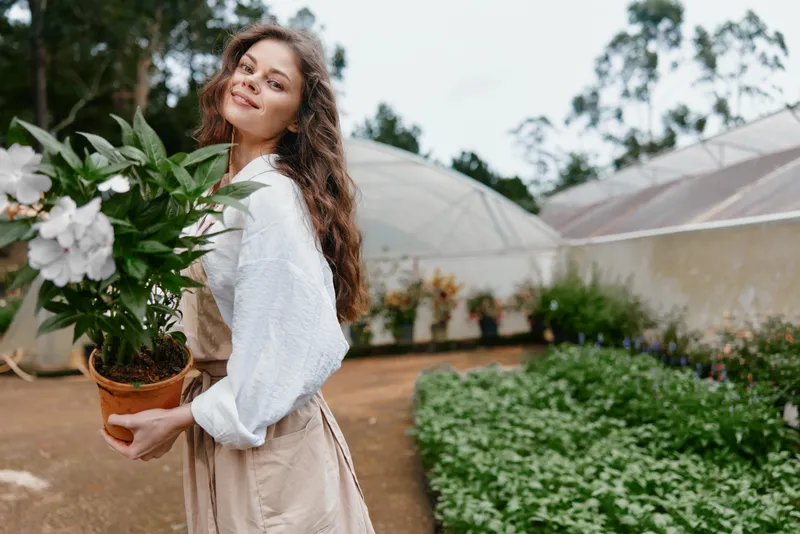Young woman with curly hair in garden holding potted flowers, joyful expression in natural setting, showcasing gardening passion, spring vibes, lush greenery